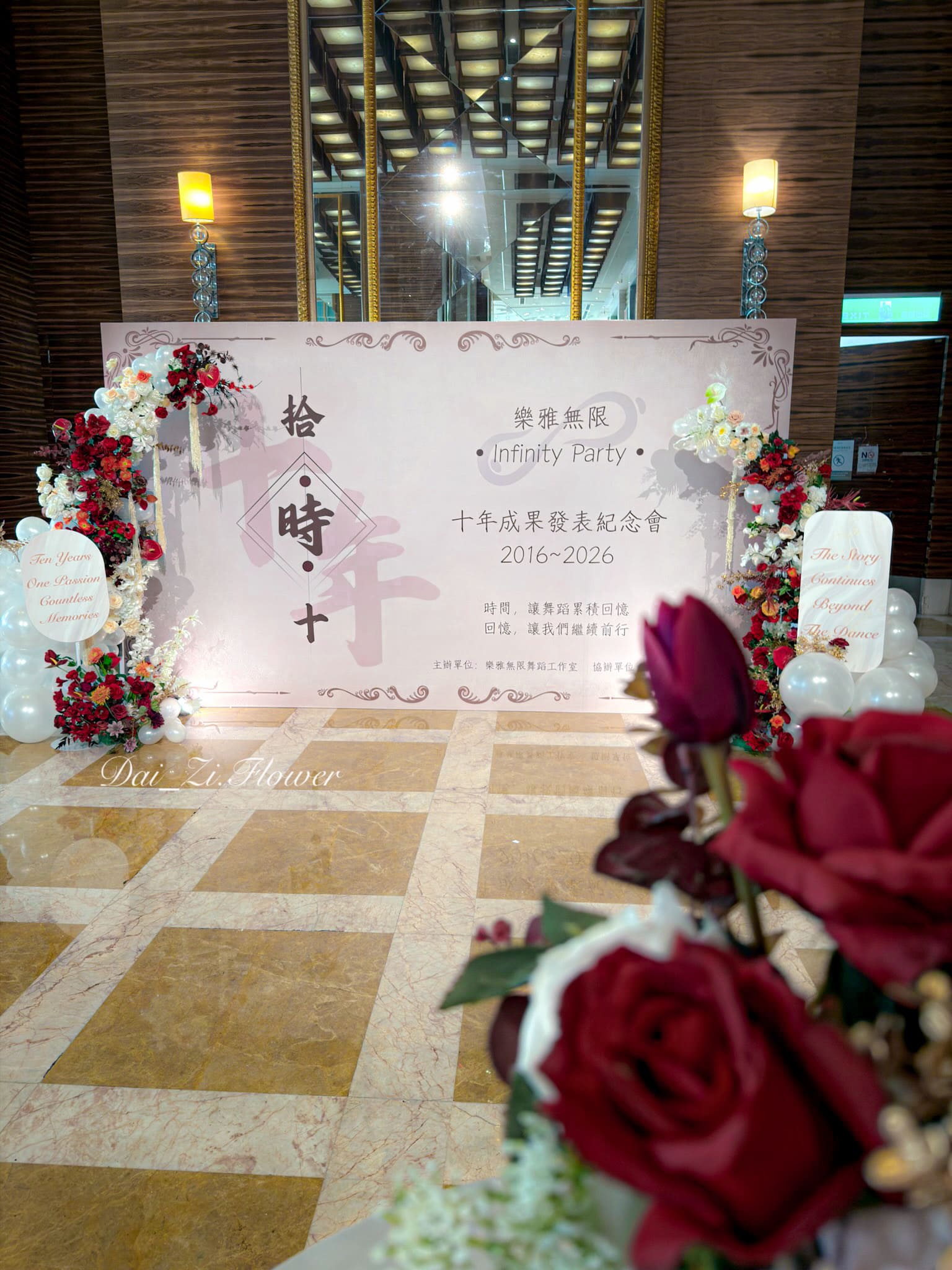 DaiZi 代紫花藝｜大直典華舞蹈十週年成果發表全場正面—粉紫色系活動背板搭配左右雙側酒紅白橘色仿真花藝柱、白色氣球群，前景紅玫瑰花束，台北典華大直宴會廳活動花藝氣球佈置